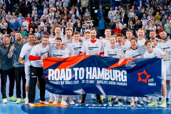 Ein jubelndes Handballteam posiert auf einem Spielfeld, lächelnd und mit einem Road to Hamburg EHF Finals-Banner in der Hand, mit einem Zebra-Maskottchen und jubelnden Fans im Hintergrund.