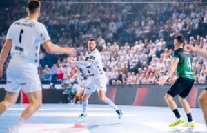 Ein Handballspieler in einem weißen Trikot bereitet sich während eines Spiels auf einen Pass vor. Zwei Gegner sind zu sehen, und im Hintergrund ist eine große, verschwommene Menschenmenge zu sehen.