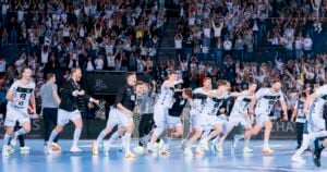 Eine Männerhandballmannschaft in weißen Trikots rennt und jubelt auf einem Hallenplatz, während eine große, jubelnde Menge auf der Tribüne hinter ihnen steht.