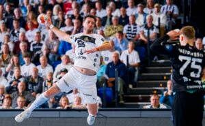 Ein männlicher Handballspieler in einem weißen Trikot springt in der Luft, um den Ball in Richtung Tor zu werfen, während der Torwart in Schwarz sich darauf vorbereitet, den Ball abzuwehren. Zuschauer beobachten das Geschehen von den Tribünen im Hintergrund.