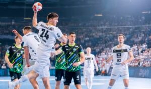 Ein Handballspieler in einem weißen Trikot springt zum Schuss, während er von Spielern in schwarzen und grünen Trikots während eines rasanten Spiels in einer überfüllten Halle verteidigt wird.
