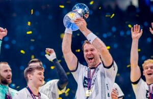 Ein jubelnder Sportler in einem weißen Trikot hebt eine glänzende Trophäe in die Höhe und lächelt, während um ihn herum Konfetti fällt. Die Mannschaftskameraden, die Medaillen tragen und jubeln, feiern fröhlich neben ihm.