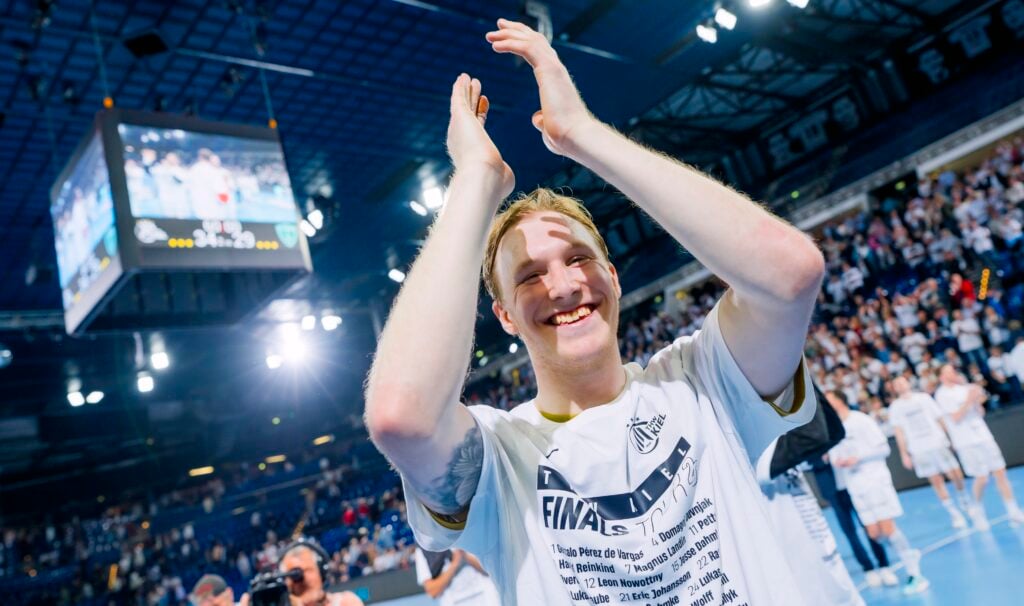 HANDBALL 2025/26: THW Kiel - RK Nexe Ein lächelnder Sportler in einem weißen Hemd klatscht in einer Sporthalle über seinem Kopf und feiert mit Fans und Teamkollegen. Ein großer Bildschirm mit seinem Bild hängt über der hellen, überfüllten Halle.