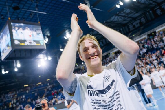 Ein lächelnder Sportler in einem weißen Hemd klatscht in einer Sporthalle über seinem Kopf und feiert mit Fans und Teamkollegen. Ein großer Bildschirm mit seinem Bild hängt über der hellen, überfüllten Halle.