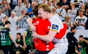 Zwei männliche Sportler in Sportuniformen umarmen sich freudig auf einem Hallenplatz, umgeben von jubelnden Zuschauern und anderen Spielern. Der eine trägt ein rotes Trikot, der andere ein weißes. Die Szene fängt die Aufregung und den Jubel ein.