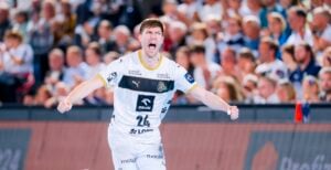 Ein männlicher Handballspieler in einem weißen Trikot feiert leidenschaftlich auf dem Spielfeld, mit ausgestreckten Armen und offenem Mund, während im Hintergrund eine Menschenmenge zusieht.
