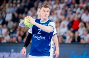 Ein männlicher Handballspieler in einem blauen Trikot mit der Nummer 2 bereitet sich darauf vor, während eines Spiels einen grünen Ball zu werfen, während ein anderer Spieler und eine unscharfe Menschenmenge im Hintergrund zu sehen sind.