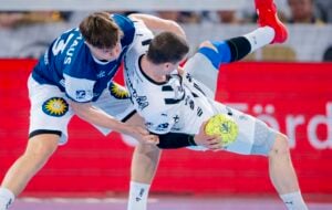 Zwei männliche Handballspieler liefern sich einen intensiven Wettkampf; der eine im blauen Trikot versucht, den Spieler im weißen Trikot, der einen gelben Ball in der Hand hält, zu blocken oder zu attackieren. Der rote Hintergrund und die Aktion deuten auf ein rasantes Spiel hin.