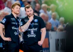 Zwei Männer in schwarzen Sportuniformen stehen nebeneinander und wirken nachdenklich, während sie ein Ereignis beobachten. Sie befinden sich in einer Reithalle mit einer unscharfen Menschenmenge im Hintergrund. Ein Mann hat seine Hand auf dem Kinn.