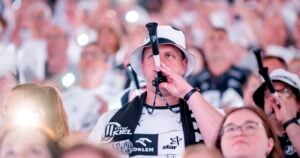 Ein Mann mit weißem Eimerhut bläst ein schwarzes Horn bei der THW Kiel EHF European League Saison 2025/2026, umgeben von Fans in Weiß und Schwarz. Einige winken mit Lichtern und tragen so zur festlichen Atmosphäre der Winamax EHF Finals Hamburg bei.