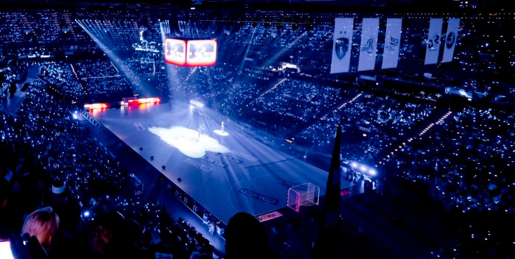 Eine große, mit Zuschauern gefüllte und mit blauem Licht beleuchtete Sporthalle. Das Spielfeld darunter ist mit Scheinwerfern und einer zentralen Videoleinwand ausgestattet, die eine dramatische Atmosphäre für eine Veranstaltung oder ein Spiel schaffen.