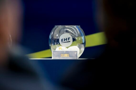Eine Glasschale mit der Aufschrift "EHF", die weiße Kugeln enthält, steht auf einem Tisch. Vor der Schale steht ein kleines Schild mit der Aufschrift "CLUBS". Der Hintergrund ist blau mit einem gelben diagonalen Streifen.
