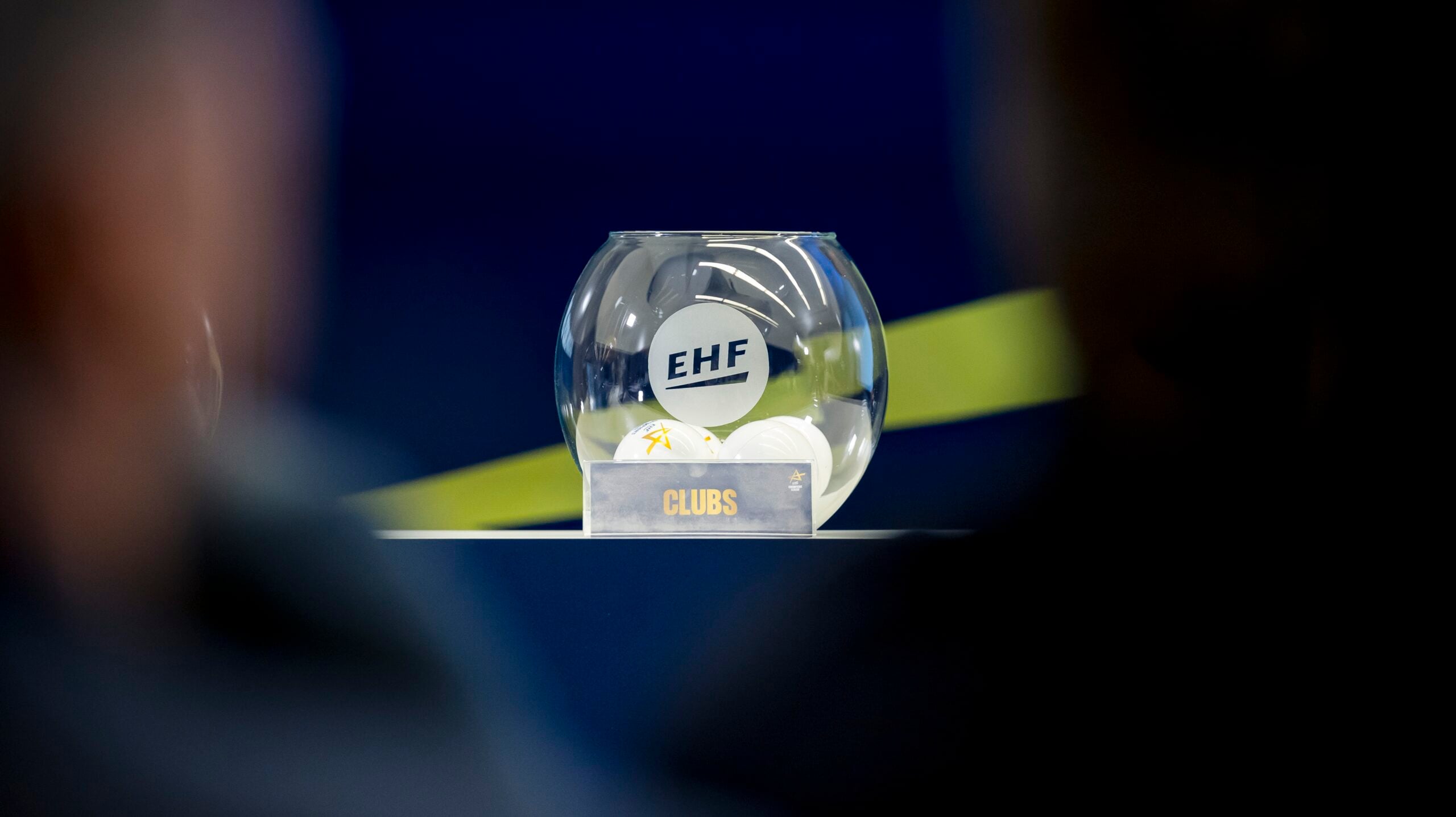 Eine Glasschale mit der Aufschrift "EHF", die weiße Kugeln enthält, steht auf einem Tisch. Vor der Schale steht ein kleines Schild mit der Aufschrift "CLUBS". Der Hintergrund ist blau mit einem gelben diagonalen Streifen.