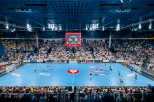 Ein großes Hallenstadion, das mit Zuschauern gefüllt ist, die ein Handballspiel auf einem blauen Spielfeld verfolgen, mit Spielern in Aktion und einer Anzeigetafel, auf der FINALE und der Spielstand angezeigt werden, bei heller Beleuchtung der Arena.