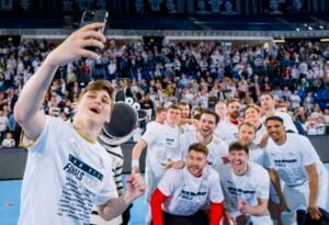Eine Gruppe lächelnder Athleten in weißen Hemden macht ein Selfie auf dem Hallenboden der Barclays Arena in Hamburg, begleitet von einem Zebra-Maskottchen. Auf den Tribünen hinter ihnen ist eine jubelnde Menge während des THW Kiel EHF Handball Final4 zu sehen.