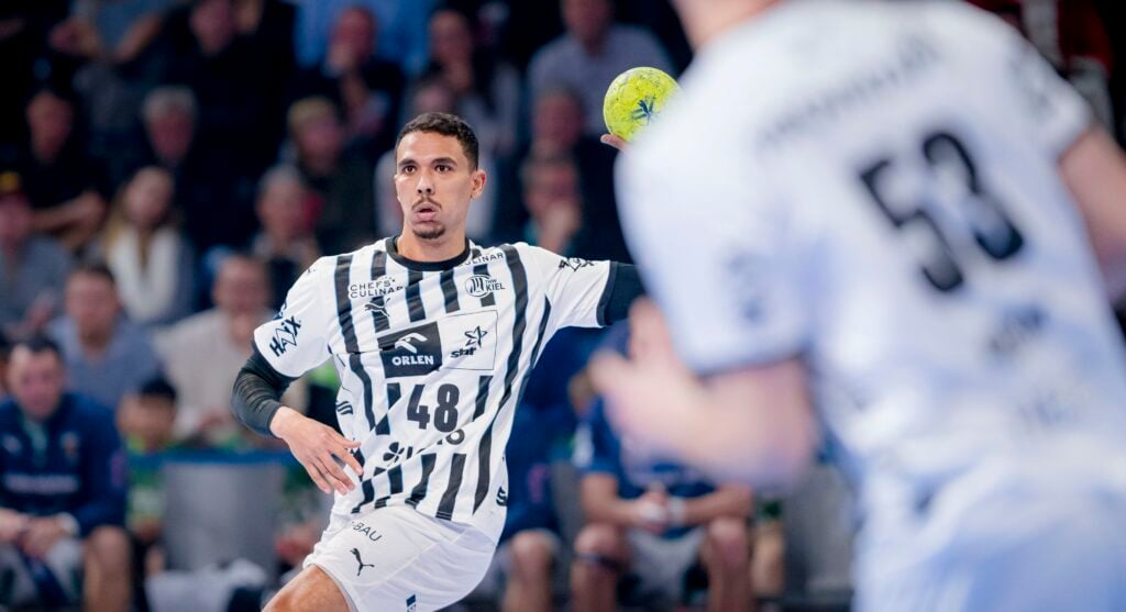 Ein Handballspieler in einem schwarz-weiß gestreiften Trikot bereitet sich auf den Wurf eines gelben Balls vor. Im Vordergrund ist ein Mitspieler zu sehen, im Hintergrund eine verschwommene Menschenmenge.