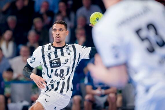Ein Handballspieler in einem schwarz-weiß gestreiften Trikot bereitet sich auf den Wurf eines gelben Balls vor. Im Vordergrund ist ein Mitspieler zu sehen, im Hintergrund eine verschwommene Menschenmenge.