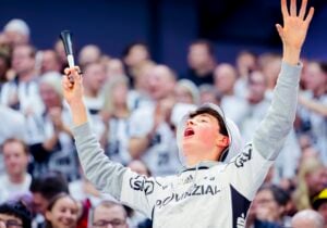 Ein junger Fan in Sportkleidung jubelt enthusiastisch mit erhobenen Armen und offenem Mund, umgeben von einer Menge unscharfer Zuschauer bei einer Hallenveranstaltung.