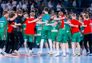 Eine Gruppe männlicher Handballspieler in grünen und roten Trikots steht in einem Kreis auf einem Hallenplatz, legt die Arme umeinander und feiert gemeinsam, während im Hintergrund eine Menschenmenge zusieht.