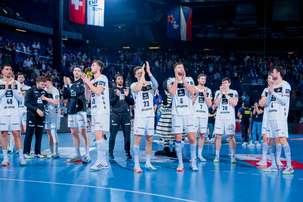 Eine Männerhandballmannschaft in weißen Trikots steht auf einem Hallenplatz, klatscht und blickt in Richtung Publikum. Die Halle ist mit Zuschauern gefüllt, und über den Tribünen im Hintergrund hängen mehrere Fahnen.