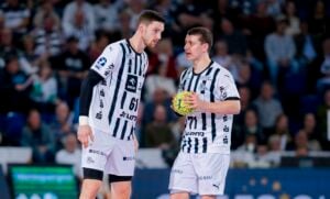 Zwei männliche Handballspieler in schwarz-weiß gestreiften Trikots stehen auf dem Spielfeld, einer hält einen gelben Ball in der Hand, und unterhalten sich während des Spiels miteinander. Im Hintergrund ist ein verschwommenes Publikum zu sehen.