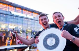 Zwei aufgeregte Männer feiern vor einer hell erleuchteten Arena, in der Hand eine große Meisterschale und ein Bier. Lächelnd markieren sie in schwarzen Trikots die Handball-Bundesliga Saison 2025/2026 des THW Kiel und den emotionalen Abschied von Petter Överby mit der jubelnden Menge im Hintergrund.