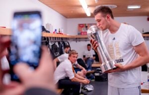 Ein Mann im weißen Hemd, der die Handball-Bundesliga-Saison 2025/2026 des THW Kiel feiert, küsst in der Umkleidekabine eine große Trophäe, während ein anderer ein Foto schießt - möglicherweise zum Abschied von Petter Överby -, während sich die Mannschaftskameraden auf den Bänken daneben ausruhen.