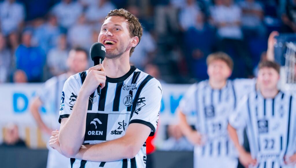 HANDBALL 2025/26: THW Kiel - TBV Lemgo Lippe Ein lächelnder Handballspieler in einem schwarz-weiß gestreiften Trikot hält ein Mikrofon und spricht auf einem Spielfeld. Im Hintergrund sind unscharfe Mannschaftskameraden in ähnlichen Trikots zu sehen.