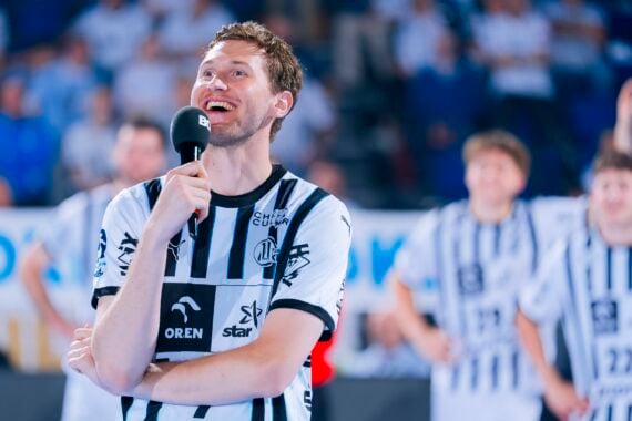 Ein lächelnder Handballspieler in einem schwarz-weiß gestreiften Trikot hält ein Mikrofon und spricht auf einem Spielfeld. Im Hintergrund sind unscharfe Mannschaftskameraden in ähnlichen Trikots zu sehen.