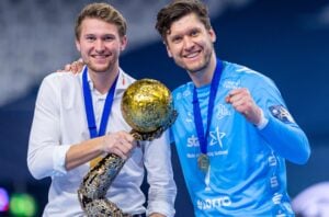 Zwei lächelnde Männer mit Medaillen posieren zusammen; einer hält eine große goldene Trophäe. Ein Mann trägt ein weißes Hemd, der andere ein blaues Sporttrikot. Im Hintergrund ist eine Sporthalle zu sehen.