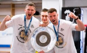 Drei junge Männer in weißen Hemden und mit Medaillen in der Hand feiern eine große silberne Trophäenplatte. Sie lächeln und scheinen aufgeregt zu sein und stehen dicht beieinander in einer hellen Halle.