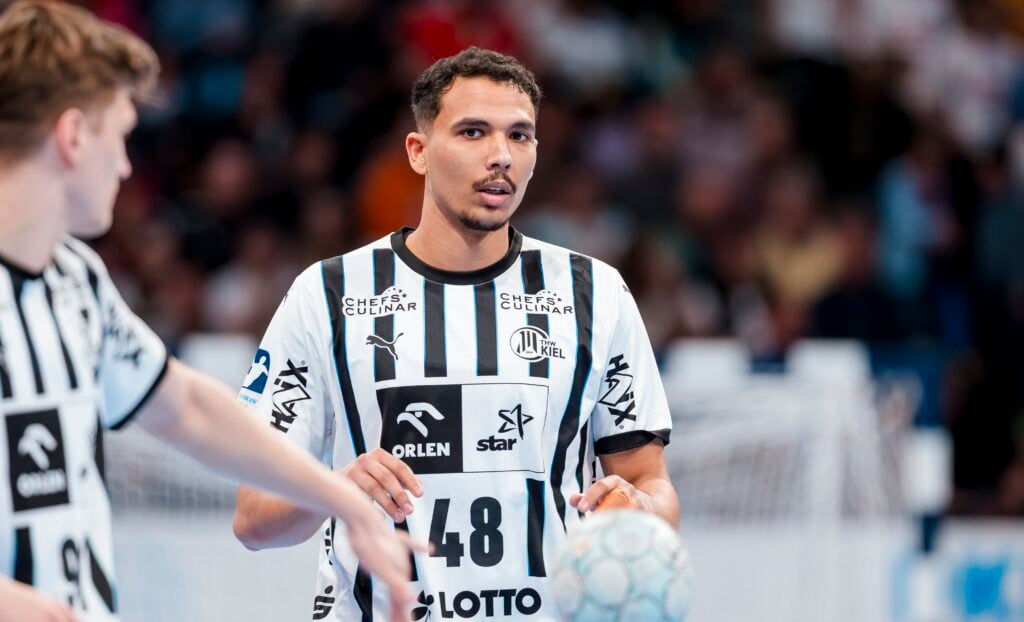 Ein männlicher Handballspieler im schwarz-weiß gestreiften Trikot des THW Kiel hält während eines Spiels der Handball-Bundesliga Saison 2025/2026 einen Ball, während sein Mannschaftskamerad Petter Överby in der Nähe ist und im Hintergrund eine unscharfe Menschenmenge zu sehen ist.