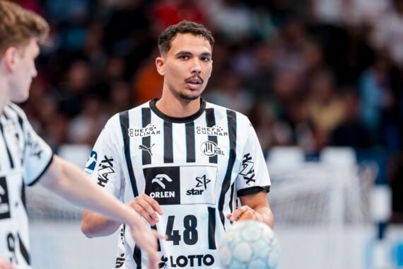 Ein männlicher Handballspieler im schwarz-weiß gestreiften Trikot des THW Kiel hält während eines Spiels der Handball-Bundesliga Saison 2025/2026 einen Ball, während sein Mannschaftskamerad Petter Överby in der Nähe ist und im Hintergrund eine unscharfe Menschenmenge zu sehen ist.