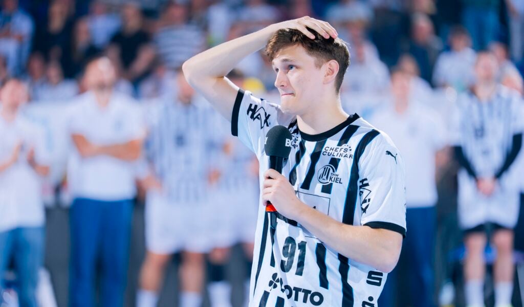 Ein junger männlicher Handballspieler in einem schwarz-weiß gestreiften Trikot, möglicherweise vom THW Kiel, steht auf dem Spielfeld, hält ein Mikrofon in der Hand und schaut nachdenklich - vielleicht zum Abschied von Petter Överby während der Handball-Bundesliga Saison 2025/2026.