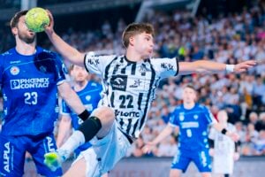 Ein Handballspieler in einem schwarz-weiß gestreiften Trikot springt hoch, um den Ball zu werfen, während ein Verteidiger in einem blauen Trikot versucht, ihn zu blockieren – all dies während eines spannenden Spiels, das von einer Zuschauermenge im Hintergrund verfolgt wird.