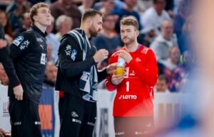 Drei männliche Sportler in Sportbekleidung stehen während einer Pause bei einer Hallenveranstaltung beieinander. Einer in einem roten Trikot hält eine Trinkflasche in der Hand, während die anderen in dunklen Trikots sich unterhalten. Im unscharfen Hintergrund schaut eine Menschenmenge zu.