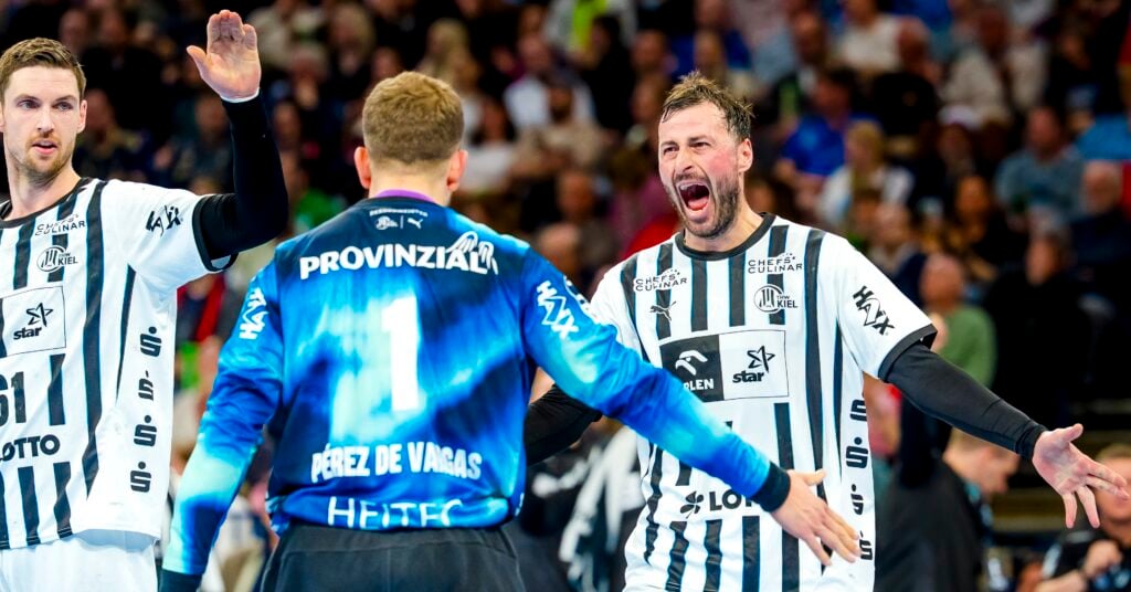 HANDBALL 2025/26: THW Kiel - HSV Hamburg Zwei Handballspieler in weißen und schwarzen Trikots feiern während eines Spiels ausgelassen mit einem Mitspieler in einem blauen Trikot, während im Hintergrund eine jubelnde Zuschauermenge zu sehen ist.