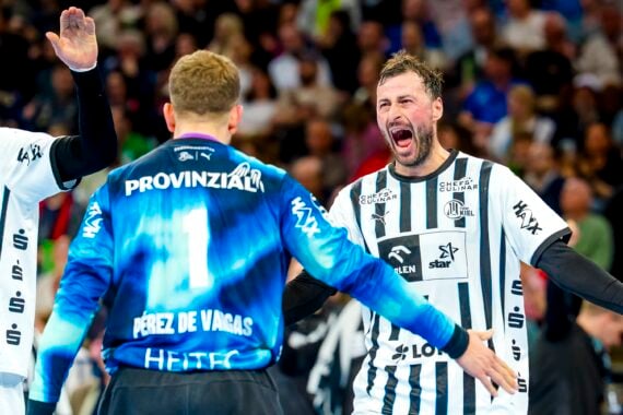 Zwei Handballspieler in weißen und schwarzen Trikots feiern während eines Spiels ausgelassen mit einem Mitspieler in einem blauen Trikot, während im Hintergrund eine jubelnde Zuschauermenge zu sehen ist.