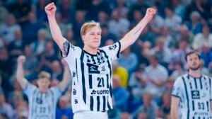 Ein Handballspieler in einem schwarz-weiß gestreiften Trikot mit der Nummer 21 reckt jubelnd beide Arme in die Höhe. Im unscharfen Hintergrund sind seine Mannschaftskameraden und eine jubelnde Menge zu sehen.