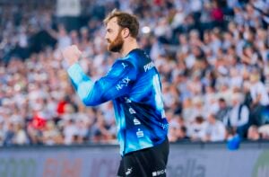 Ein Handballspieler in einem blau-schwarzen Trikot reckt jubelnd die Faust in die Höhe. Er blickt mit konzentriertem Gesichtsausdruck nach rechts, im Hintergrund ist eine verschwommene Menschenmenge zu sehen.