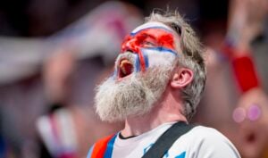 Ein Mann mit grauem Bart und rot-weiß-blauer Gesichtsbemalung jubelt begeistert beim Wenzel Nordic Cup im Rahmen der Vorbereitung des THW Kiel auf die Handball-Bundesliga-Saison 2026/2027, im Hintergrund sind verschwommene Fans auf den Färöern zu sehen.
