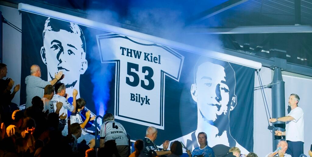 Fans applaudieren vor einem großen Trikotbanner mit der Aufschrift THW Kiel 53 Bilyk. Zwei Graustufenporträts flankieren das Trikot, während im Hintergrund blauer Rauch zu sehen ist.