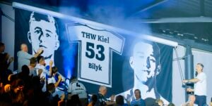 Fans applaudieren vor einem großen Trikotbanner mit der Aufschrift THW Kiel 53 Bilyk. Zwei Graustufenporträts flankieren das Trikot, während im Hintergrund blauer Rauch zu sehen ist.