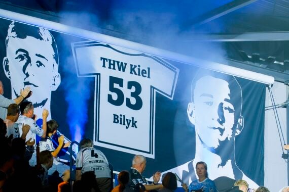 Fans applaudieren vor einem großen Trikotbanner mit der Aufschrift THW Kiel 53 Bilyk. Zwei Graustufenporträts flankieren das Trikot, während im Hintergrund blauer Rauch zu sehen ist.