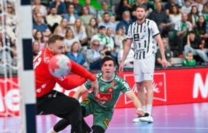 Ein Handballtorwart in Rot versucht, einen Schuss zu blocken, während ein Spieler in Grün zu Boden fällt. Ein anderer Spieler in Schwarz und Weiß schaut im Hintergrund zu, während auf der Tribüne eine Menge Zuschauer sitzt.