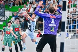 Ein Handballspieler in einem weißen Trikot springt, um den Ball in Richtung Tor zu werfen, während sich zwei Verteidiger in Grün und der Torwart in Lila darauf vorbereiten, ihn abzuwehren. Die Zuschauer schauen von den Tribünen im Hintergrund zu.