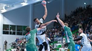 Ein Handballspieler in einem weiß-schwarzen Trikot springt, um den Ball zu werfen, während zwei Verteidiger in grünen Trikots versuchen, ihn zu blockieren. Die Zuschauer verfolgen das Geschehen von der Tribüne aus in einer hell erleuchteten Halle.