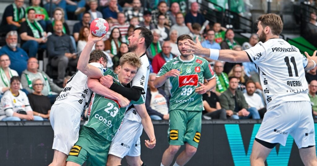 Ein spannender Moment während eines Handballspiels zeigt, wie ein Spieler in Grün von mehreren Gegnern in weißen Trikots verteidigt wird, während die Zuschauer auf der Tribüne aufmerksam zusehen.