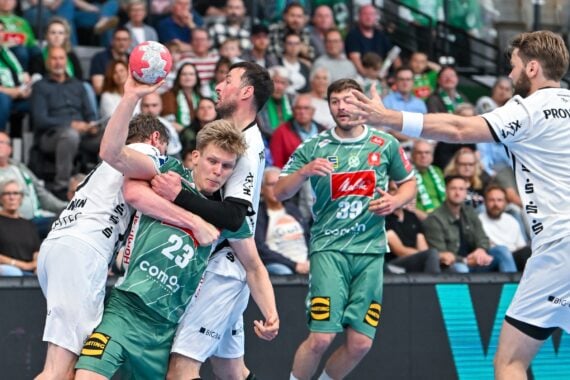 Ein spannender Moment während eines Handballspiels zeigt, wie ein Spieler in Grün von mehreren Gegnern in weißen Trikots verteidigt wird, während die Zuschauer auf der Tribüne aufmerksam zusehen.