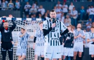 Ein männlicher Handballspieler in einem schwarz-weiß gestreiften Trikot hält ein Mikrofon in der Hand und wendet sich an ein Publikum. Mannschaftskameraden in ähnlichen Uniformen stehen hinter ihm auf einem Spielfeld, im Hintergrund sind ein Netz und Zuschauer zu sehen.
